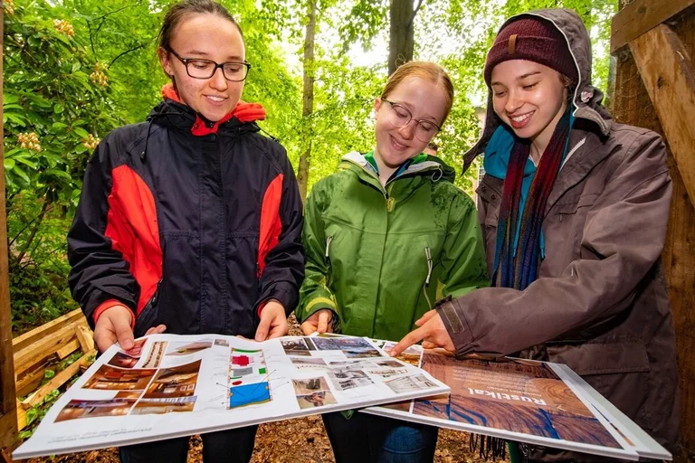 Drei junge Frauen schauen sich Pläne und Fotos an. Sie stehen im freien, unter grünen Bäumen. 