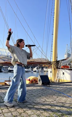 Eine junge Frau steht vor einem Segelschiff und singt.