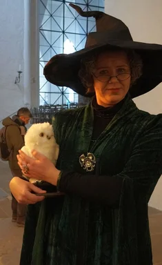 Eine Frau mit dunklen Gewand und spitzem Hexenhut hält eine Stoffeule in der Hand.