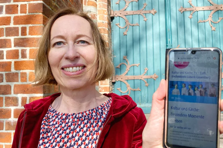 Eine Frau blickt lächelnd in die Kamera und hält ein Smartphone in die Kamera.