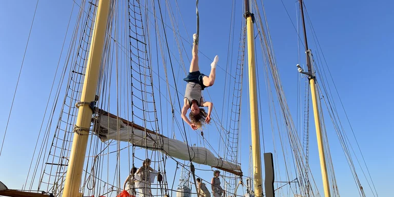 Eine junge Artistin hängt an einem Seil vor der Kulisse eines Segelschiffs.