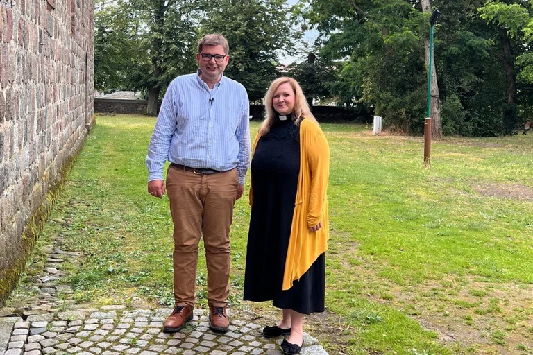 Pastorin Poepping und Pastor Gienke stehen vor einer Mauer und lächeln in die Kamera.