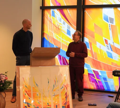 Zwei Männer stehen sich gegenüber vor einer bunt gestalteten Fensterfront.